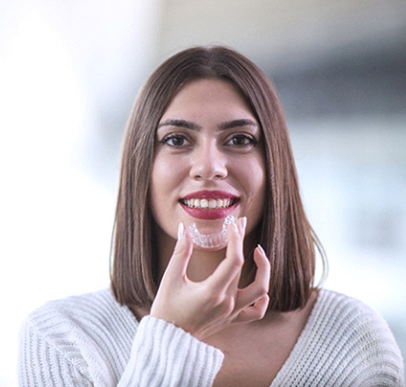 A woman wearing a white sweater holds an Invisalign aligner in preparation of reinserting it into her mouth